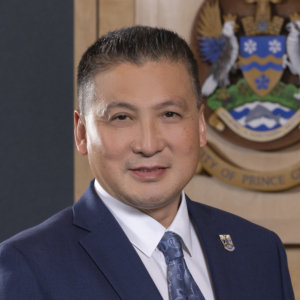 A photo of Mayor Yu wearing a navy blue suit jacket, white shirt, and blue tie with a paisley pattern. Mayor Yu wears a lapel pin with the City of Prince George Coat of Arms, and stands in front of a wall-hung version of the Coat of Arms.